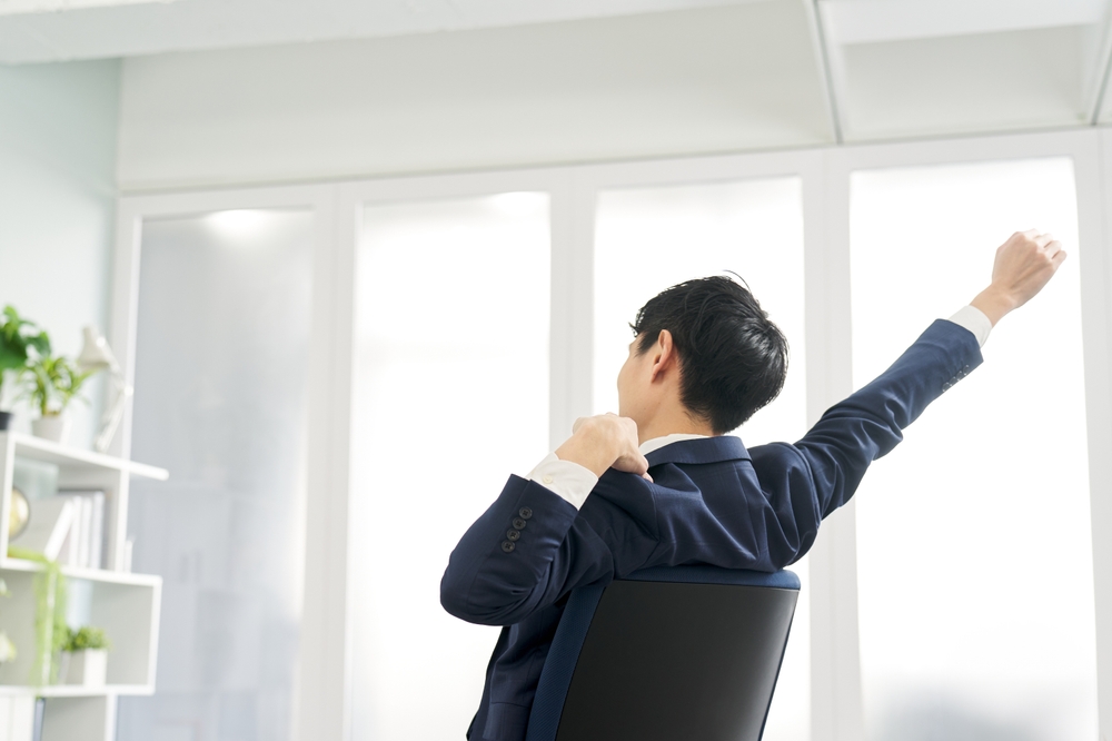デスクで伸びをするスーツの男性の後ろ姿