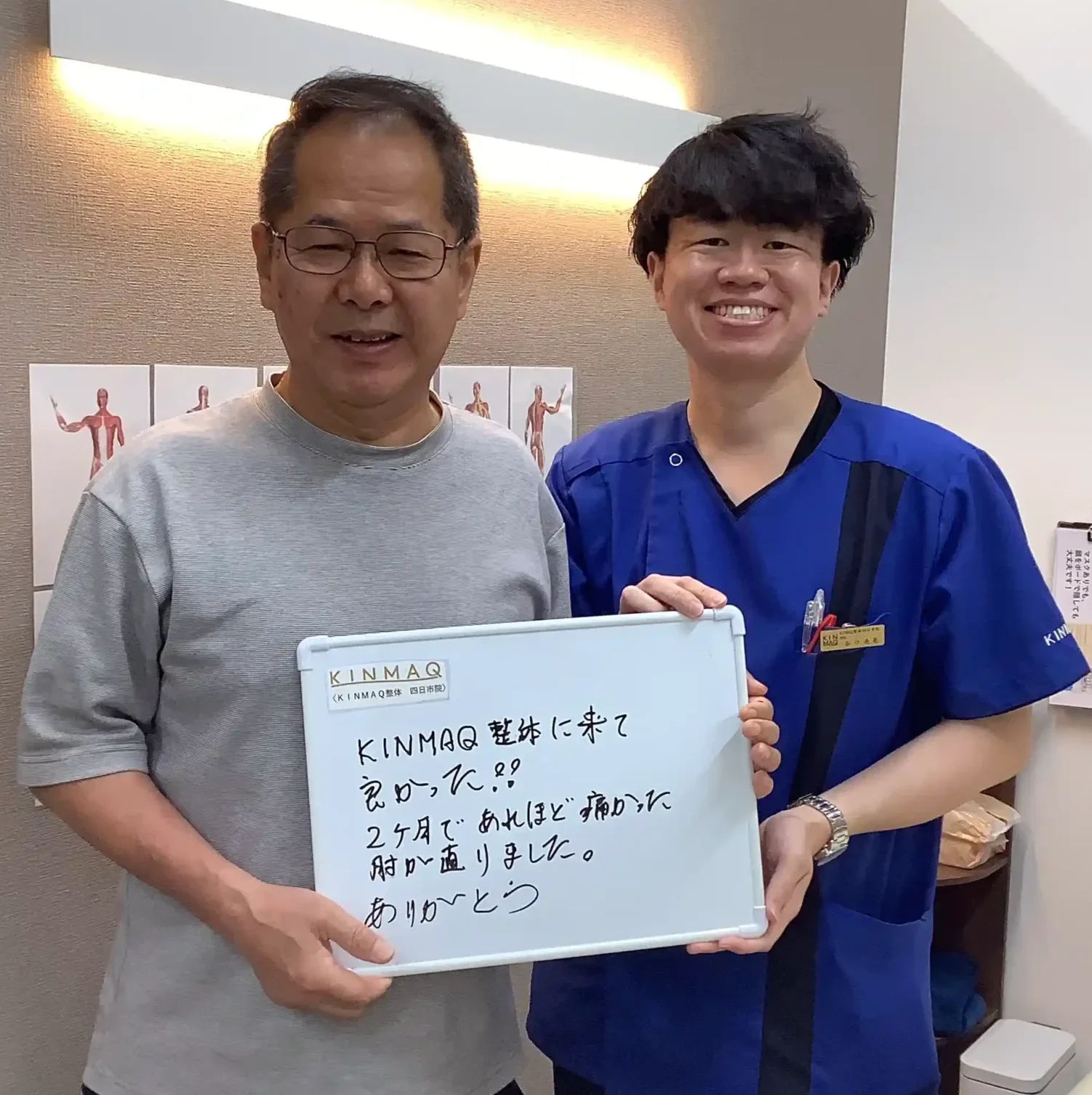 四日市の整体院で肘の痛みの改善のために通院していた男性の改善実績として。施術後にご本人が『KINMAQ整体に来て良かった！2ヶ月であれほど痛かった肘が直りました。ありがとう』と書いたホワイトボードを持ち、担当した理学療法士と2ショットを撮影したときの写真。